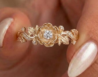 May include: A gold ring featuring a central round brilliant-cut diamond set within a sculpted floral motif. The band is designed with intricate, textured leaf patterns that extend outward from the flower, accented by small embedded diamonds.