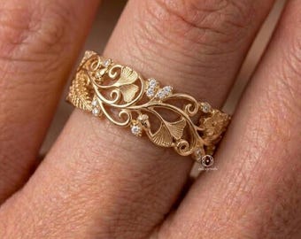 May include: A gold band ring featuring an intricate openwork design of swirling filigree patterns and ginkgo leaf motifs, accented with small, clear, round-cut stones embedded throughout the metal structure.