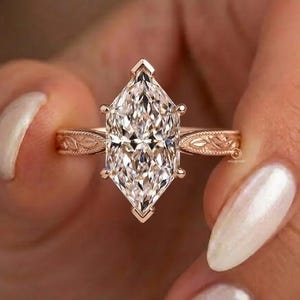 May include: A rose gold ring featuring a large marquise-cut centre stone with a faceted surface. The band is crafted from rose gold and displays an intricate, engraved leaf-like pattern along its entire outer surface, extending from the setting.