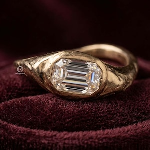 May include: A gold ring featuring a central emerald-cut clear gemstone set within a textured, organic-shaped metal band. The metal surface displays a hammered, uneven finish, and a small logo is visible on the side of the ring setting.
