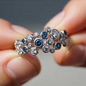 Op de afbeelding: Een zilveren ring met een cluster van ronde edelstenen. De ring heeft een uniek ontwerp met een mix van heldere en diepblauwe stenen, wat een sprankelend effect creëert. De edelstenen zijn gezet in een zilveren band.
