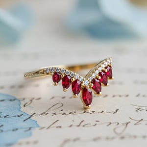 May include: A gold-colored ring with a V-shaped design. The ring features a row of small, clear gemstones and a series of larger, teardrop-shaped red gemstones. The brand name "OnicaJewels" is visible on the ring's band. The ring is set against a backdrop of handwritten script.
