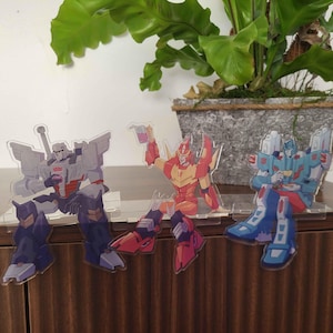 May include: Three acrylic character standees featuring stylized robot figures in seated poses. The left figure is grey and purple, the center figure is red and orange holding a drink, and the right figure is blue and white, each on a clear base.