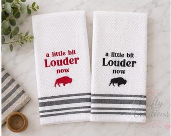 May include: Two white waffle-weave kitchen towels featuring the text a little bit Louder now and a buffalo silhouette. One towel has red text and a red buffalo, while the other has black text and a black buffalo. Both have grey striped borders.