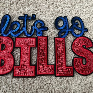 May include: A decorative patch featuring the text "let's go" in blue sequins and the word "BILLS" in red sequins. The letters are outlined in black, creating a bold, high-contrast design with a shimmering, textured surface throughout the entire piece.