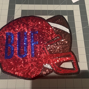 May include: A sequined patch shaped like a football helmet in red, white, and brown. The helmet features the letters BUF in blue sequins on the side, a white stripe detail, and a dark brown face mask area, all outlined in a dark border.