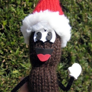 Puede incluir: Un juguete de punto marrón que lleva un gorro de Papá Noel rojo y blanco con ojos negros y boca roja. El juguete tiene guantes blancos y brazos de limpiapipas negros.