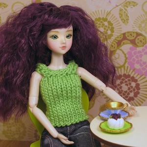 May include: A doll with long, dark purple hair wearing a green knitted tank top and blue jeans. The doll is sitting on a green chair and holding a small white cake with a gold rim on a blue and white saucer.