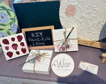 May include: A display of DIY paint kits and finished art pieces. The kits include small canvases with embossed designs and paintbrushes. Finished art includes a strawberry design and floral textured paintings. A sign reads "DIY Paint Kits".