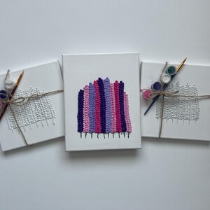May include: Three white canvas art pieces with textured designs. One canvas features a colorful, vertical striped pattern in pink, purple, and magenta. The other two canvases have a textured, woven design. Each canvas is bundled with paint and a paintbrush.