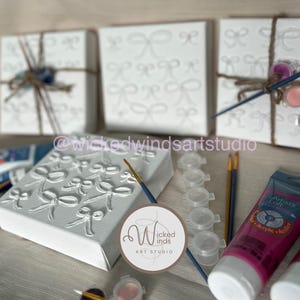May include: White canvas art kits with raised bow designs, paint pots, brushes, and acrylic paint tubes. The kits are wrapped with twine and decorative elements. The image includes the text "@wickedwindsartstudio".