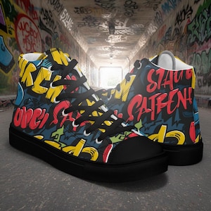 Puede incluir: Zapatillas altas con suela y cordones negros. Las partes superiores de lona están cubiertas de arte graffiti colorido, con letras rojas, amarillas y azules que dicen "Stay Patient" y "Open". Los zapatos están sobre un fondo cubierto de graffiti.