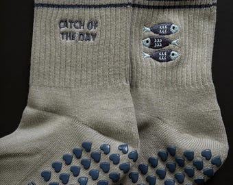 Puede incluir: Un par de calcetines grises con textura acanalada. La banda superior tiene dos rayas azul marino. La frase "CATCH OF THE DAY" está bordada en gris, junto con tres peces azules. Las suelas tienen agarres en forma de corazón azul marino.
