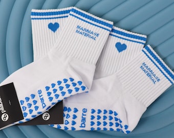Puede incluir: Un par de calcetines blancos con rayas azules en los puños, un icono de corazón azul y el texto "MARRIAGE MATERIAL" bordado en azul. Las suelas tienen agarres de silicona en forma de corazón y la palabra "barre" en color azul.