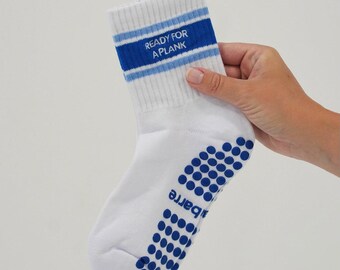 Puede incluir: Un calcetín blanco de media caña con dos rayas horizontales azules en el puño y el texto READY FOR A PLANK bordado en azul. La suela del calcetín está cubierta con puntos de agarre circulares azules y la palabra barre impresa en azul.