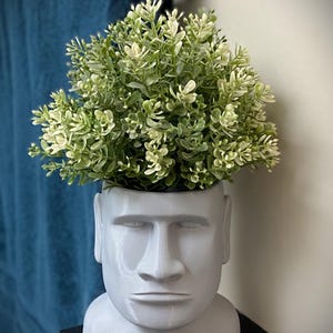May include: A gray, Moai head-shaped planter filled with green and white faux foliage. The planter has a smooth finish and is placed on a black surface. The background includes a blue towel and a silver fixture.