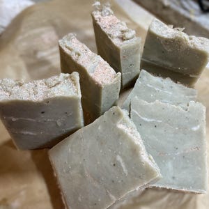 May include: A collection of handmade soap bars in various shapes and sizes. The soaps have a light green hue with visible flecks and a textured top. Some bars have a pinkish layer on top. The soaps are arranged on a brown paper surface.