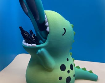 May include: A green dinosaur-shaped desk organizer with a wide-open mouth holding several black pens. The figure features a gradient color transition from light green to dark green, black circular spots on its side, and a row of small spikes.