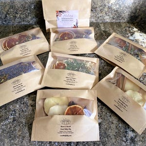 May include: An assortment of brown paper bags with clear windows, each containing dried botanicals and spices. Some bags feature dried orange slices, while others contain herbs and cinnamon sticks. The bags are labeled with text and a logo.
