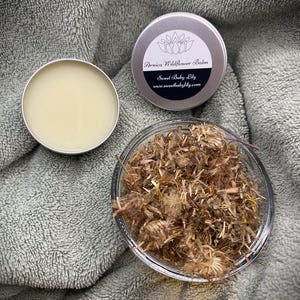 以下が含まれることがあります： 乾燥ハーブの瓶、小さな開いたバーム缶、そして「Arnica Wildflower Balm Sweet Baby Lily www.sweetbabylily.com」のテキストが入った閉じた缶のクローズアップ。灰色のタオルに置かれています。