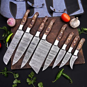 May include: A set of eight kitchen knives with patterned steel blades and wooden handles. The knives are arranged on a wooden cutting board, with a blue leather knife roll in the background. Vegetables such as tomatoes, garlic, and peppers are also visible.