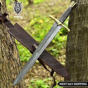 May include: A Viking sword with a patterned blade and a gold-coloured crossguard. The handle is wrapped in a dark material. The sword is in a brown leather sheath. The image includes the text "NEXT DAY SHIPPING".