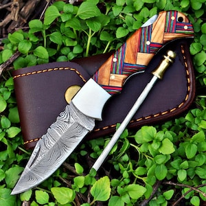 May include: A folding knife with a patterned wooden handle featuring red, blue, and green accents. The blade has a unique etched design. It rests on a brown leather sheath with a sharpening rod.