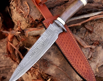 May include: A Damascus steel knife with a brown handle and a white accent, resting in a brown leather sheath. The knife blade has a unique pattern. The sheath has a stitched design and a leather strap with a metal button.