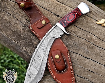 May include: A Damascus steel hunting knife with a red and black wooden handle and a brown leather sheath. The blade has a unique pattern. The sheath has a belt loop and snap closure. The knife is displayed on a wooden surface.