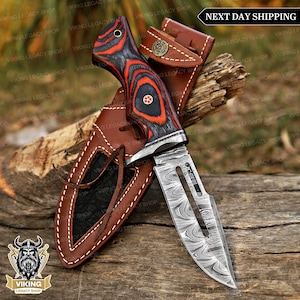 May include: A Damascus steel knife with a red and black patterned handle, in a brown leather sheath. The blade has a decorative design, displayed on wood. The text "NEXT DAY SHIPPING" is visible. The knife is approximately 20 cm long.