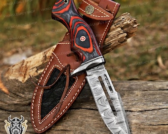 May include: A Damascus steel knife with a red and black patterned handle, resting in a brown leather sheath. The knife has a decorative design on the blade and is displayed on a wooden surface. The text "NEXT DAY SHIPPING" is visible.