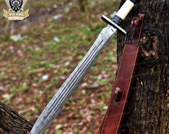 May include: A double-edged sword with a patterned Damascus steel blade, a crossguard, and a handle featuring a cream-colored grip with black accents. The sword is accompanied by a brown leather sheath with a strap and a metal button closure.