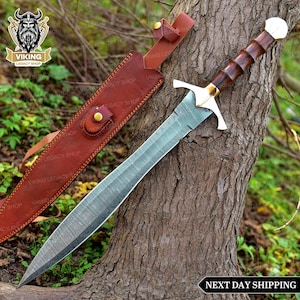 May include: A Viking sword with a patterned steel blade, a wooden handle, and a gold-coloured crossguard. The sword is displayed with a brown leather sheath. The image includes the text "NEXT DAY SHIPPING".