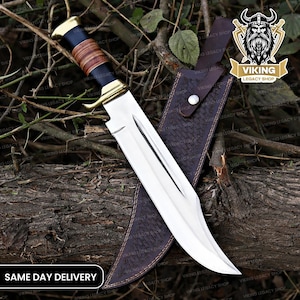 May include: A large, polished steel knife with a curved blade, brass accents, and a black and brown handle. It rests in a dark brown leather sheath. The image includes the text "SAME DAY DELIVERY" and a logo with a Viking helmet.