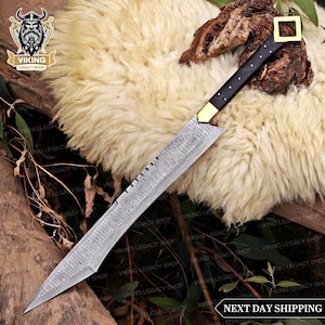 May include: A Viking-style sword with a patterned blade and a black handle with gold accents. The sword has a square gold guard and a pointed tip. The blade has a serrated edge near the handle. The sword is displayed on a fluffy, cream-coloured surface.