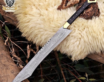 May include: A Viking-style sword with a patterned blade and a black handle with gold accents. The sword has a square gold guard and a pointed tip. The blade has a serrated edge near the handle. The sword is displayed on a fluffy, cream-colored surface.