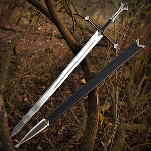 May include: A long, silver sword with a black handle and crossguard, in a black sheath. The sword features an ornate hilt with decorative details. The background is a blurred view of a wooded area with brown branches.