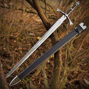 May include: A long, silver sword with a detailed blade and ornate crossguard, next to a black sheath. The hilt is black with silver detailing. The sword is set against a backdrop of tree branches and foliage.