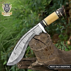 May include: A Damascus steel knife with a curved blade and a bone handle. The handle is cream-coloured with black and gold accents. The knife is resting on a piece of wood. The blade has a unique pattern.