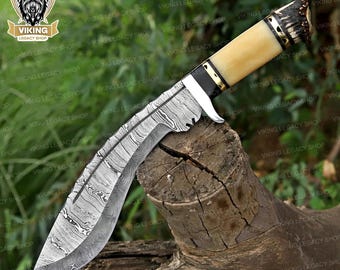 May include: A Damascus steel knife with a curved blade and a bone handle. The handle is cream-colored with black and gold accents. The knife is resting on a piece of wood. The blade has a unique pattern.