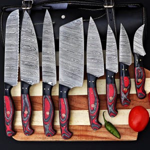 Op de afbeelding: Een set van acht Damascus stalen keukenmessen met rood-zwarte houten handvatten. De messen hebben een uniek wervelpatroon. De messen liggen op een houten snijplank met een tomaat en een groene chilipeper.
