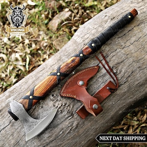 Può includere: Un'ascia vichinga con manico in legno, dettagli in pelle nera e lama argentata. Viene mostrata con un fodero in pelle marrone. L'immagine include il testo "VIKING LEGACY SHOP" e "NEXT DAY SHIPPING".