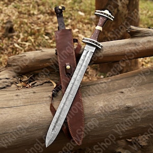 May include: A sword with a patterned blade and a wooden handle with white accents, displayed with its brown leather sheath. The sword rests on a weathered wooden log. The blade has a detailed pattern. The sheath is brown leather.