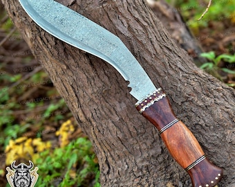 May include: A large, curved knife with a patterned blade and a wooden handle. The handle is dark brown with brass accents. The blade has a textured appearance. The knife is leaning against a tree trunk.
