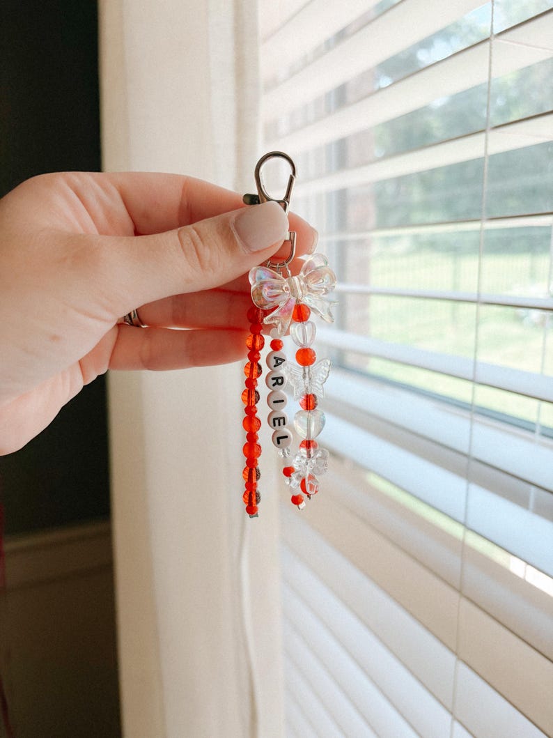 May include: A beaded keychain featuring a silver-toned lobster clasp, a translucent iridescent bow, and three dangling strands. One strand contains red beads, another features white letter beads spelling ARIEL, and the third has clear butterfly beads.