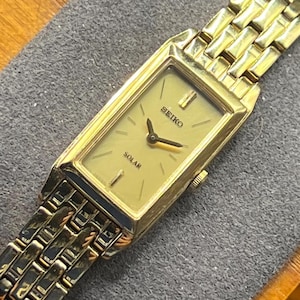 May include: A gold-toned Seiko Solar watch with a rectangular face and a matching gold link band. The watch face is a light gold color with black hands and the word "SEIKO" and "SOLAR" printed on it. The watch is displayed on a gray surface.