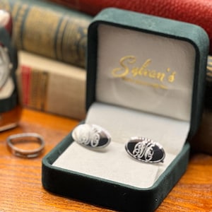 May include: Oval cufflinks with a black and silver design, displayed in a green velvet box. The box is open, revealing the cufflinks with an engraved monogram. The box is labeled "Sylvan's". A ring and a watch are also visible.