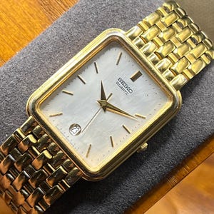 May include: A gold-toned Seiko Quartz watch with a square face and a gold link band. The watch face is white with gold hands and markers, and a date display at the 3 o'clock position. The watch is displayed on a gray surface.