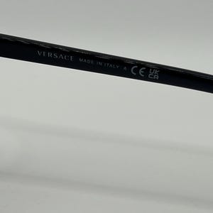 Peut inclure: Gros plan sur une monture de lunettes noire avec le nom de la marque "VERSACE" et "MADE IN ITALY" imprim&eacute;s sur la branche. La monture porte &eacute;galement un marquage "CE" et les symboles "UK CA".