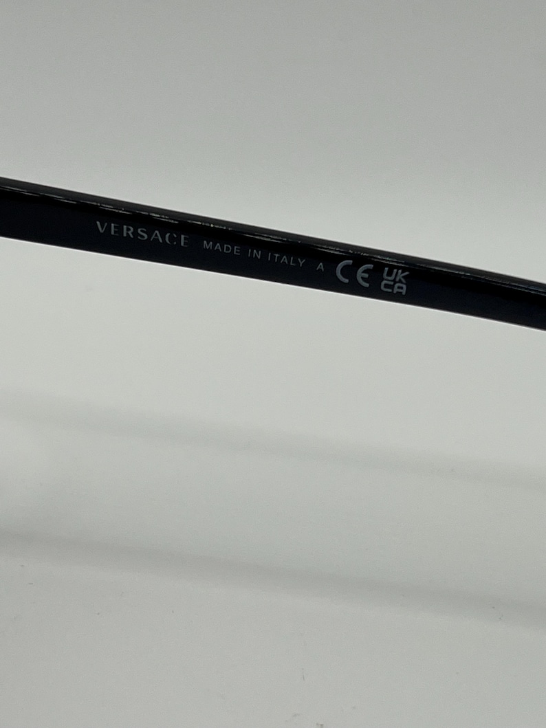 Peut inclure: Gros plan sur une monture de lunettes noire avec le nom de la marque "VERSACE" et "MADE IN ITALY" imprim&eacute;s sur la branche. La monture porte &eacute;galement un marquage "CE" et les symboles "UK CA".